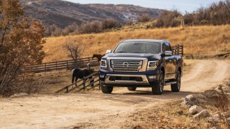 A 2022 Nissan Titan pickup in dark blue passes some horses on a dirt road. The truck has a large chrome grille, chrome side mirror caps, and chrome running boards.