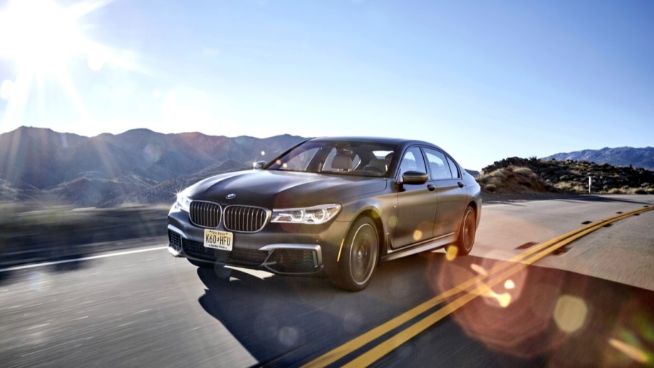 The 2022 BMW 7 Series drives on a mountain road. The car is dark gray, long, and low. We see it from a front quarter angle, facing to our left.