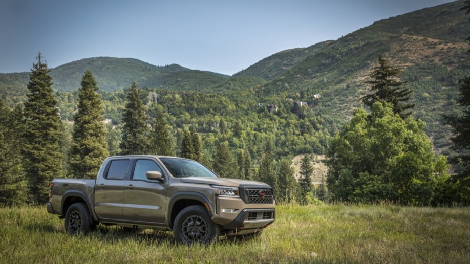 A 2022 Nissan Frontier pickup sits parked in a grassy field. The truck is a muted green color. We see it from a front quarter angle, facing to our right,