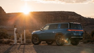 A Rivian R1S electric SUV sits plugged into a charger in a gravel parking lot against a canyon backdrop. The SUV is medium blue. We see it from a rear quarter angle.