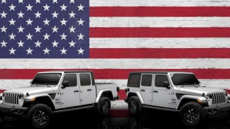The 2023 Jeep Wrangler and Jeep Gladiator Freedom Editions are parked against an American flag backdrop. Both are silver with small black and white American flag decals ahead of the doors. The SUV faces right, the truck left.