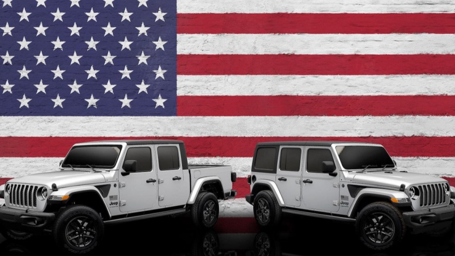 The 2023 Jeep Wrangler and Jeep Gladiator Freedom Editions are parked against an American flag backdrop. Both are silver with small black and white American flag decals ahead of the doors. The SUV faces right, the truck left.