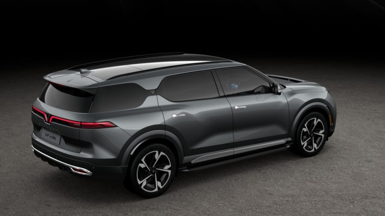 The 2023 VinFast VF9 is parked against a gray backdrop. We see it from a rear quarter angle, facing to our right. It is gunmetal gray, with long porportions and an unusual rear-facing vent stretchin the full width of its hood.