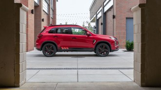 A 2022 Mitsubishi Outlander Sport sits parked between brick buildings. It is red. We see it in profile.