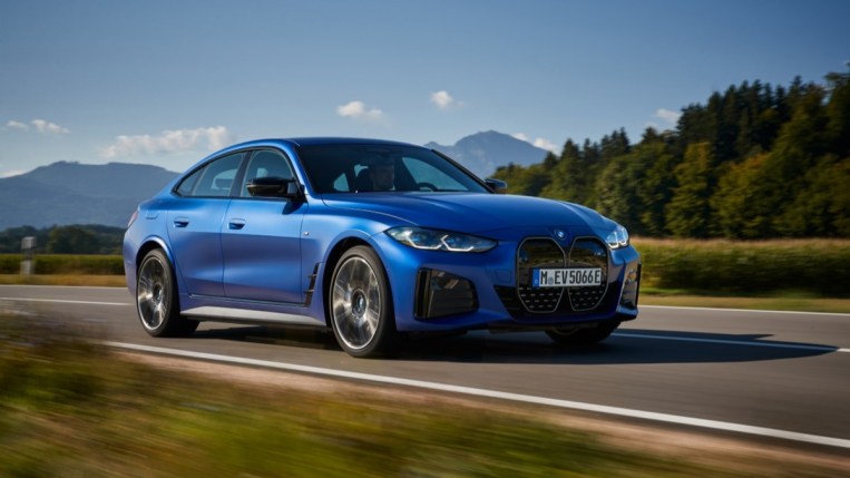 The 2023 BMW i4 electric sedan drives past a forest. The car is medium blue. We see it from a front quarter angle, facing slightly to our right. 