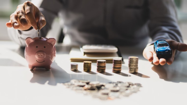 A person holds a toy car next to a piggy bank and several piles of coins of escalating size